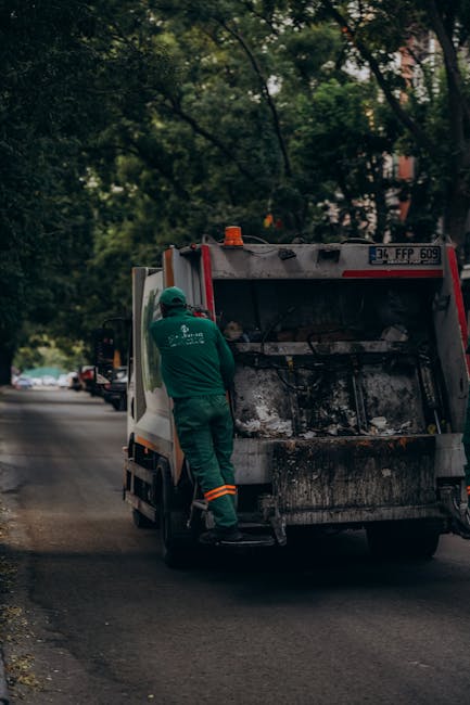 A municipal waste collection worker wearing a high-visibility orange and yellow vest is standing beside the rear of a large red rubbish truck, which is positioned on the side of a paved road. The work