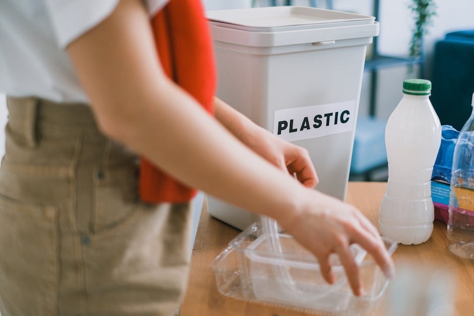 A person with long red hair, dressed in a white shirt and beige trousers, is holding a clear plastic bag in their hands, preparing for rubbish disposal. To the left, there is a white metal bin with a 