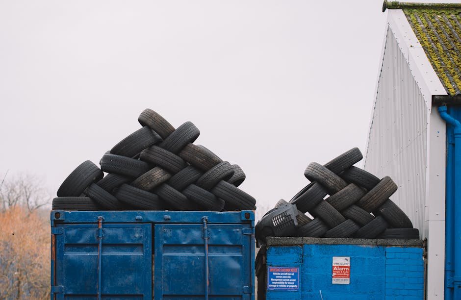 A green plastic wheeled rubbish bin placed on a paved surface next to a dark brick wall, filled with red and black flexible garden hoses or tubing coiled and layered on top of each other. The hoses ha